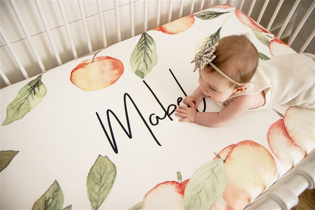 personalized crib sheet for a baby with watercolor peaches on it and the name of baby written in the middle of a peach frame that reaches all around the edges of custom crib sheet