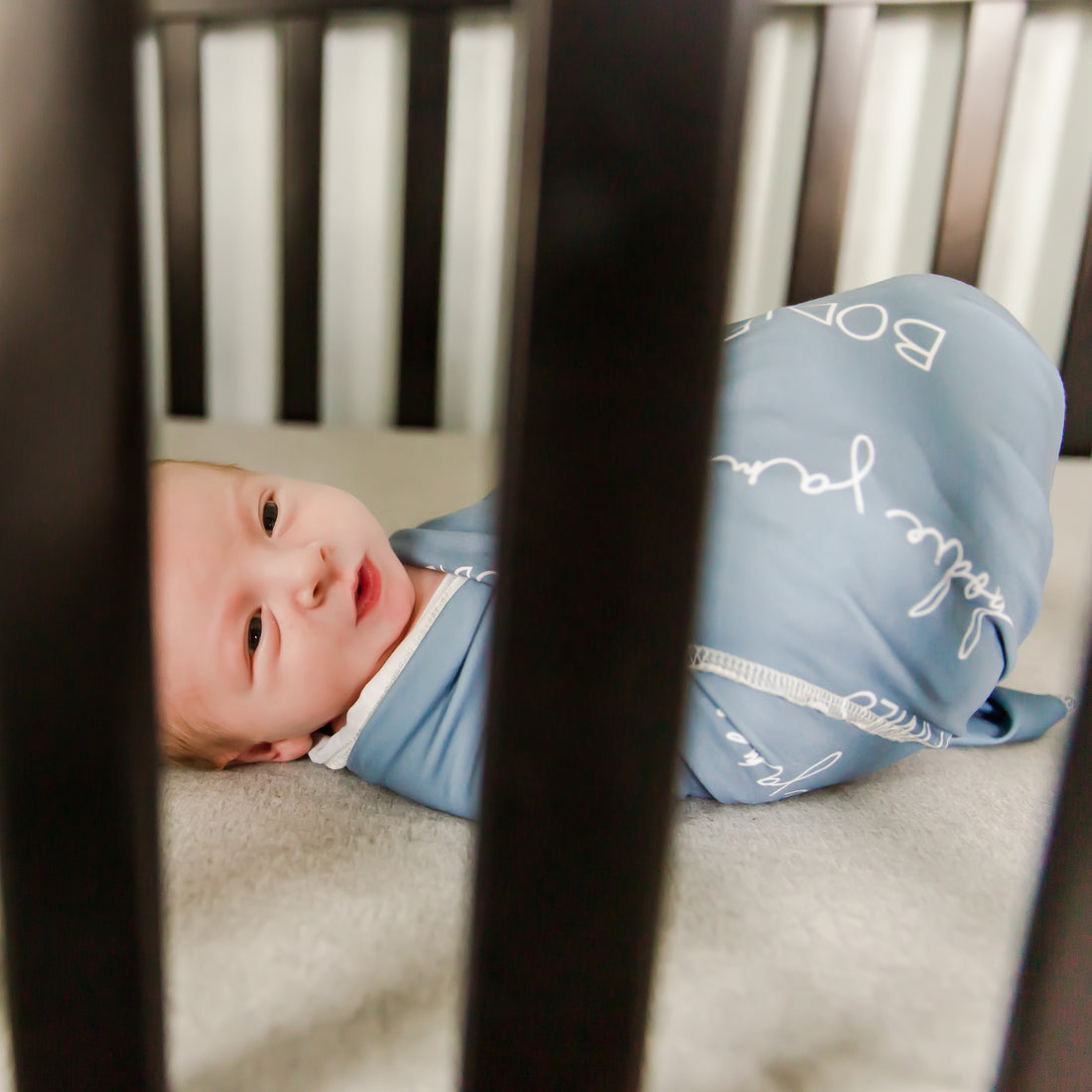A baby laying in a crib swaddled in their personalized stretchy camden blue swaddle blanket.