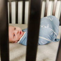 A baby laying in a crib swaddled in their personalized stretchy camden blue swaddle blanket.