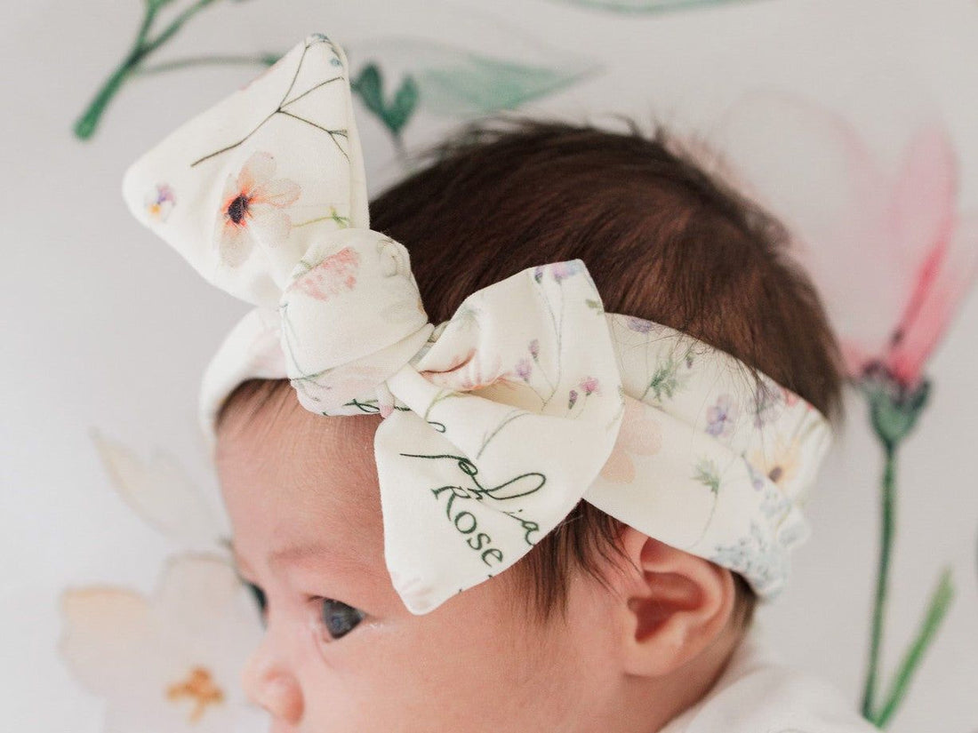 A close-up of a baby's head wearing a white floral headband with the name "Rose" printed on it.