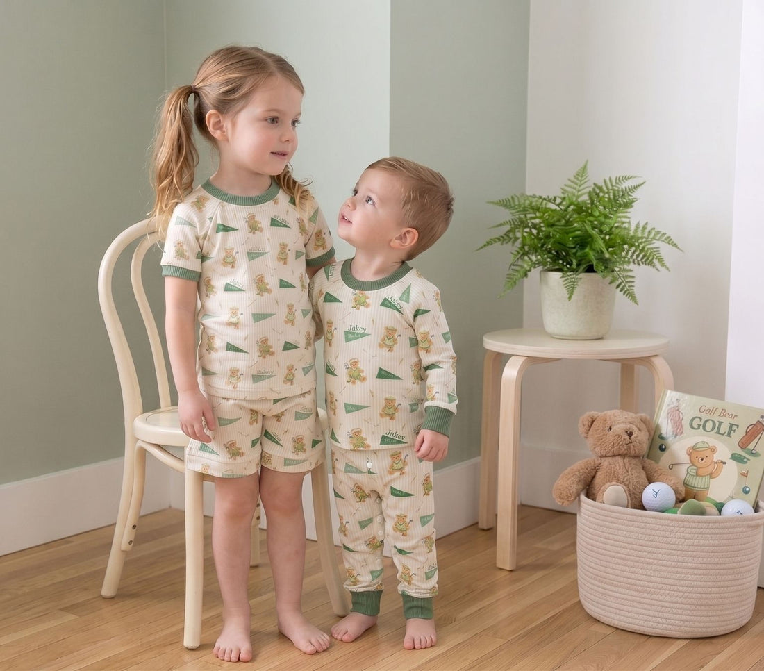 Two young children wearing matching pajamas stand together in a cozy room, with a basket of toys and a potted plant nearby.