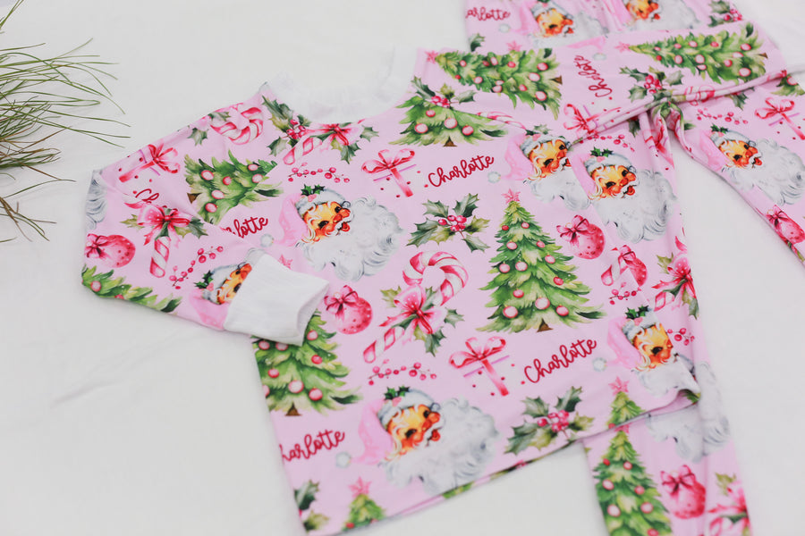 custom holiday sleepwear for little girls, with vintage santa faces and christmas trees with pink bows on a light pink material, name in red script font, and white cuffs.