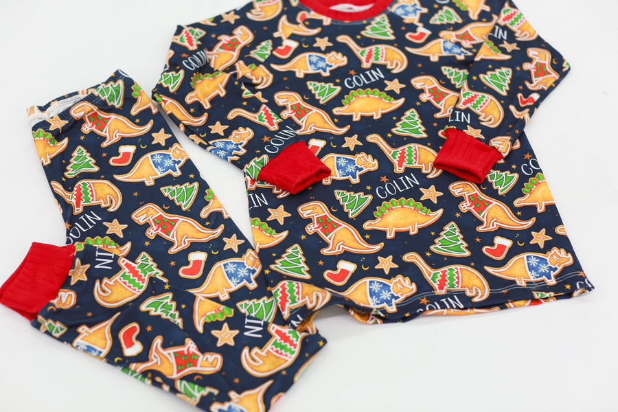 Little boy wearing personalized Dino Cookie print Christmas pajamas, navy blue background with dinosaur shaped cookies wearing Christmas sweaters