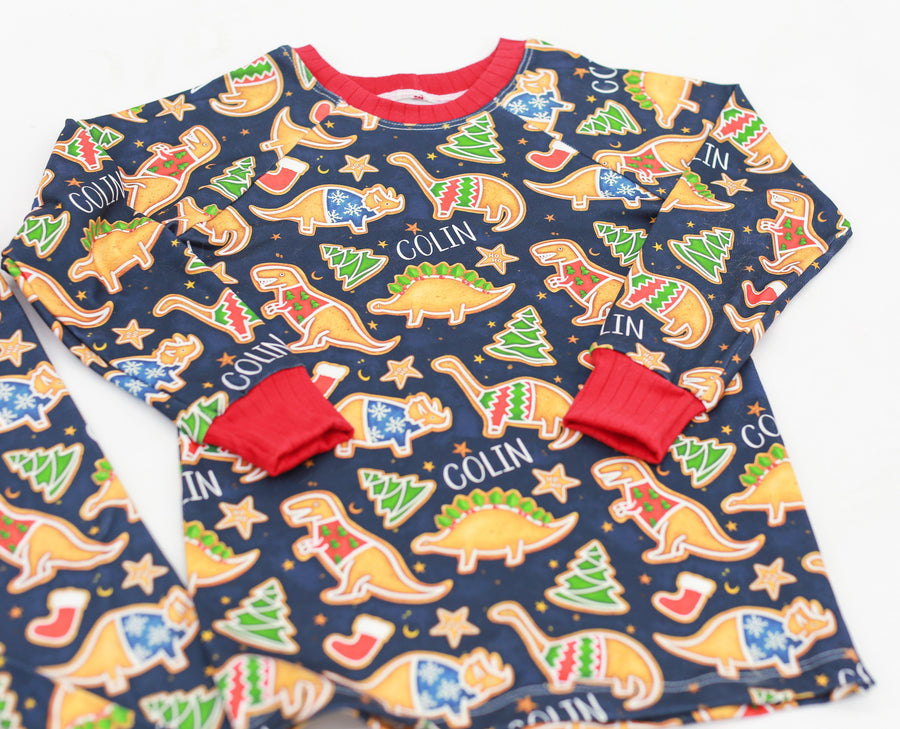 Little boy wearing personalized Dino Cookie print Christmas pajamas, navy blue background with dinosaur shaped cookies wearing Christmas sweaters