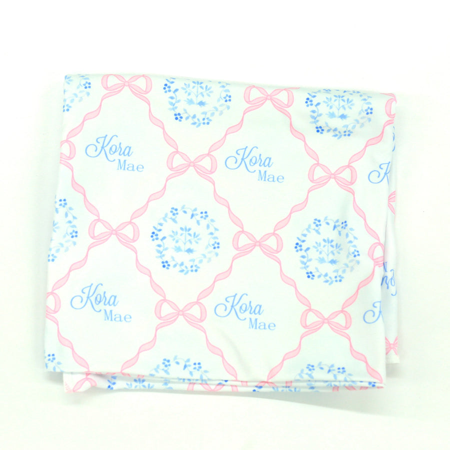  pink heirloom bow print personalized baby swaddle on white fabric and baby's name in light blue script font.