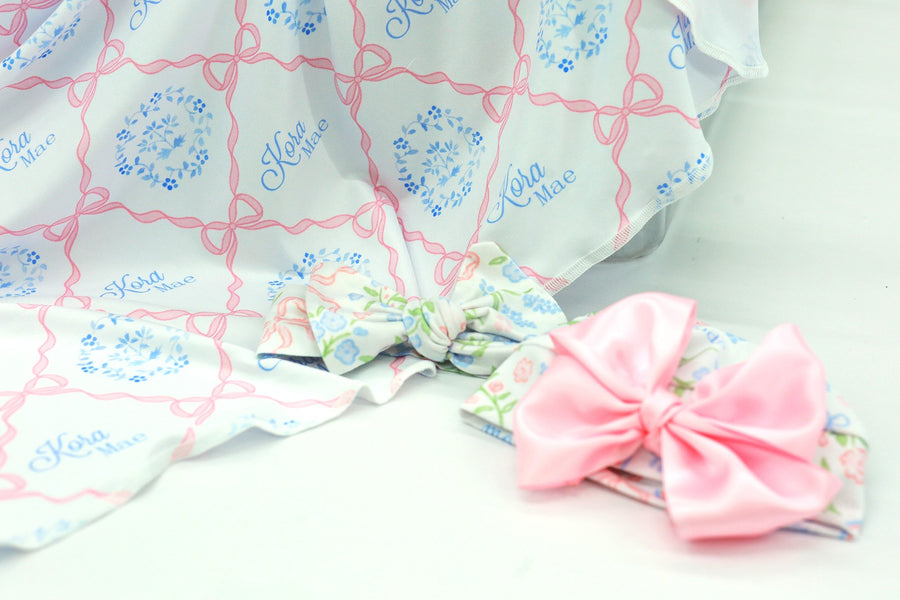 pink heirloom bow print personalized baby swaddle on white fabric and baby's name in light blue script font.