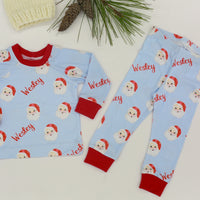 Children's pajama set with Santa Claus pattern on a light blue background, featuring the child's name and red cuffs.