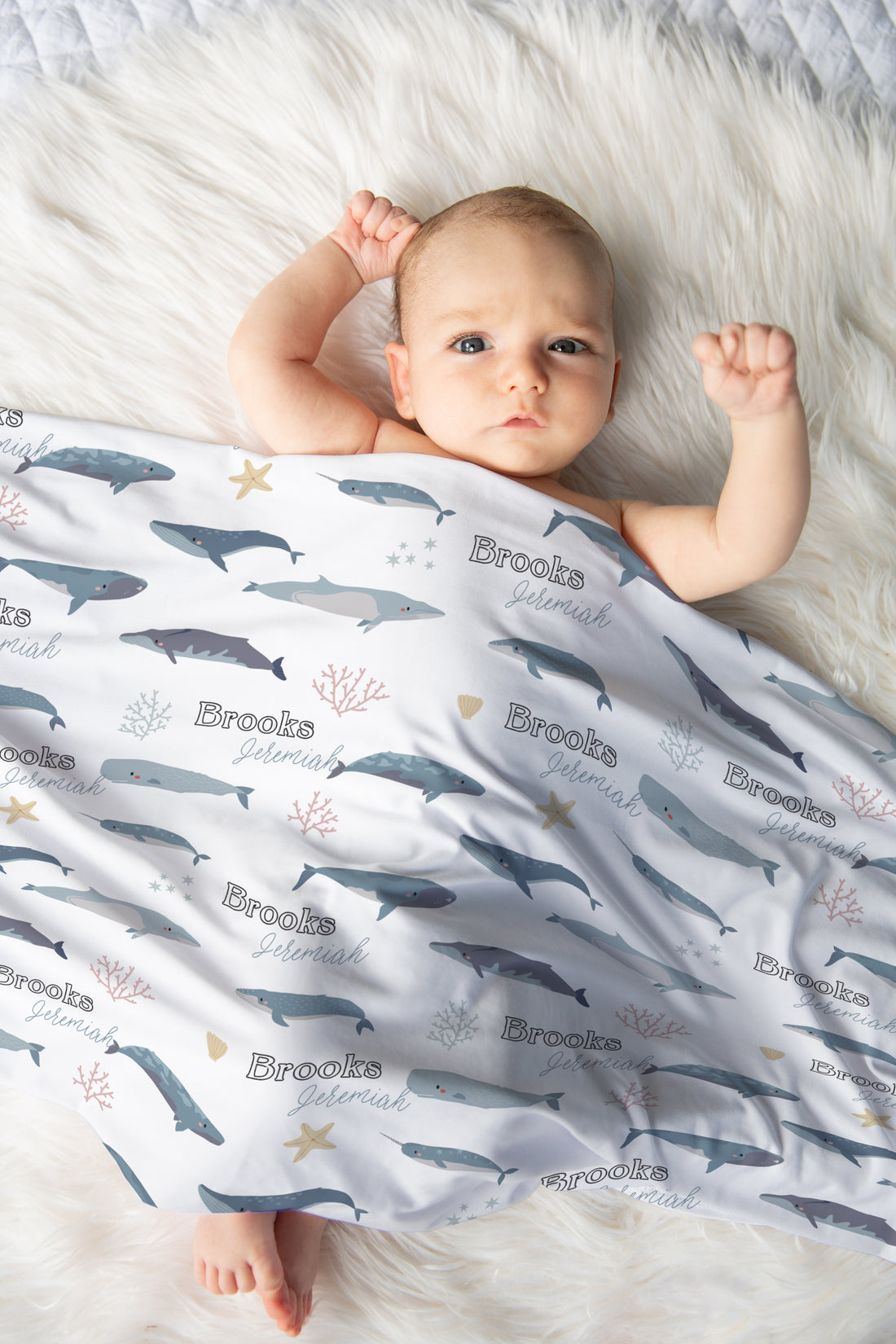 A baby is lying on a white fur-covered surface, wrapped in a blanket with a repeating pattern of blue and gray sea creatures, including sharks and starfish.