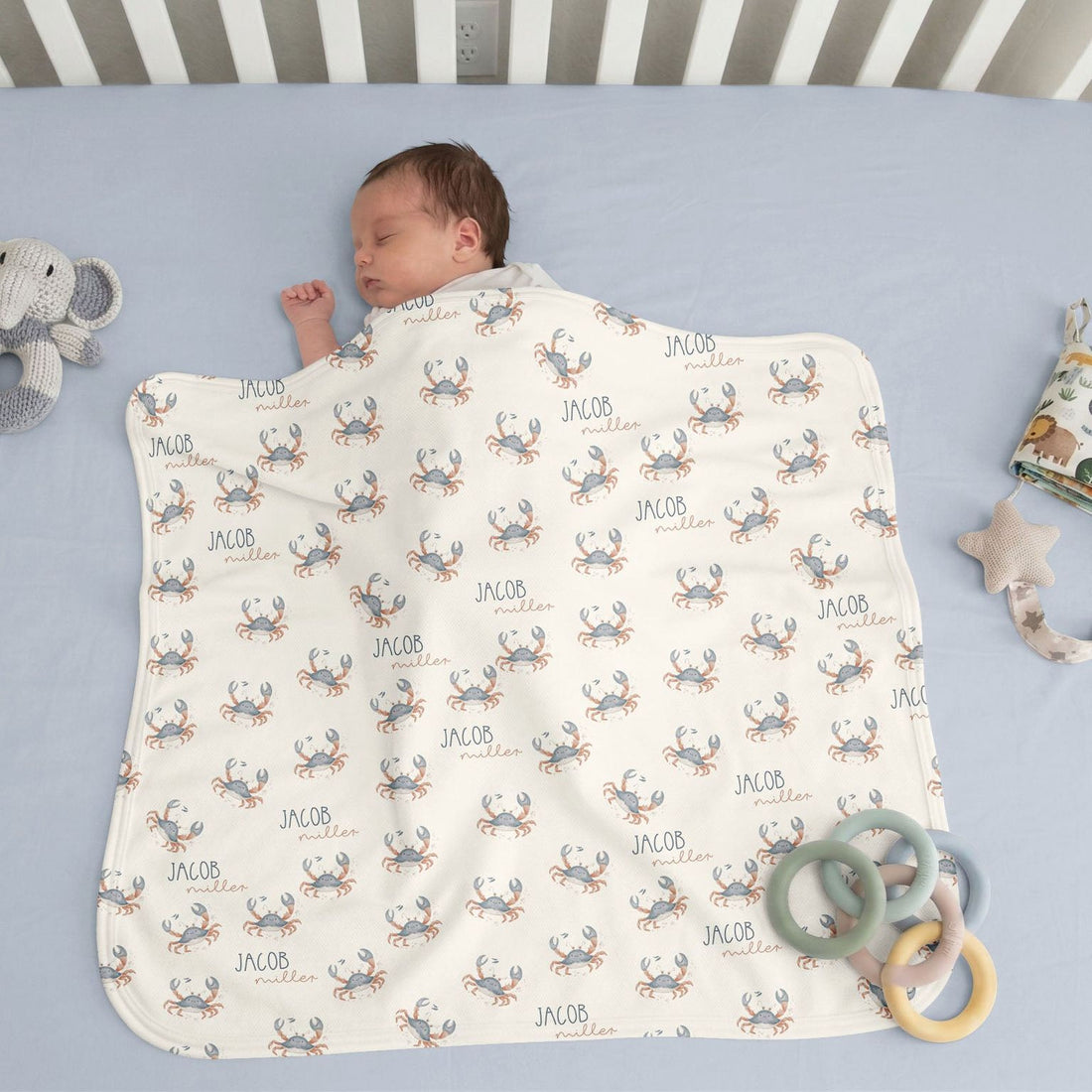 A sleeping baby wrapped in a blanket with a crab pattern, surrounded by toys on a light blue crib.