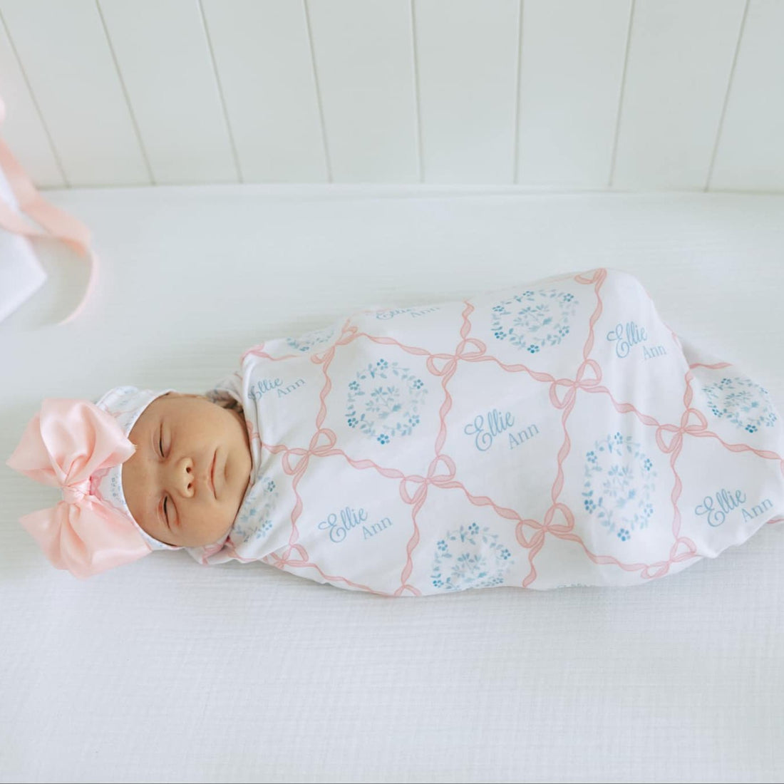 baby girl swaddled in a pink heirloom bow print personalized baby swaddle on white fabric and baby's name in light blue script font.