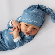 A baby wearing a blue outfit with the name "Clayton Riggs" printed on it, lying on a white background.