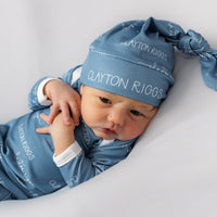 A baby wearing a blue outfit with the name "Clayton Riggs" printed on it, lying on a white background.