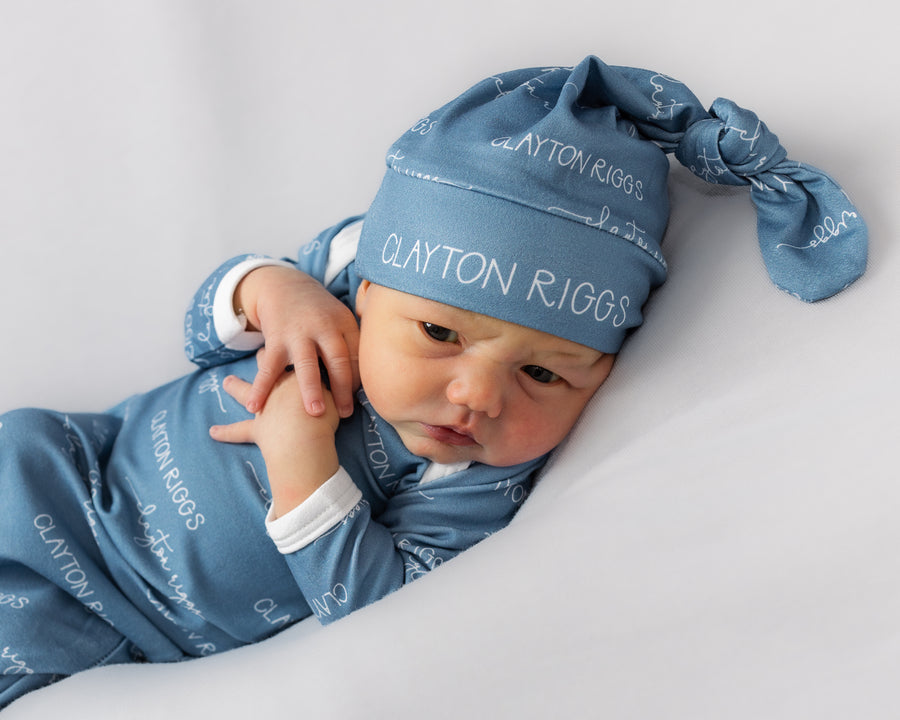 A baby wearing a blue outfit with the name "Clayton Riggs" printed on it, lying on a white background.