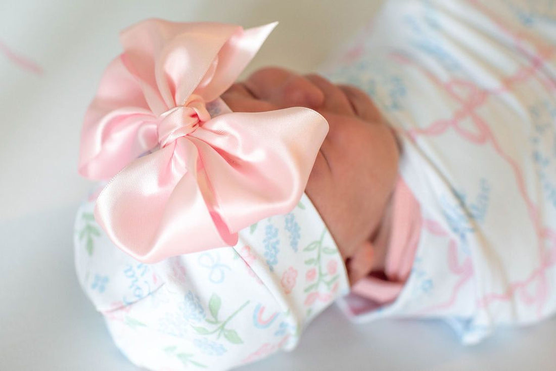 A close-up image of a baby's head wearing a pink bow on a white blanket with a floral pattern.