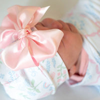 A close-up image of a baby's head wearing a pink bow on a white blanket with a floral pattern.