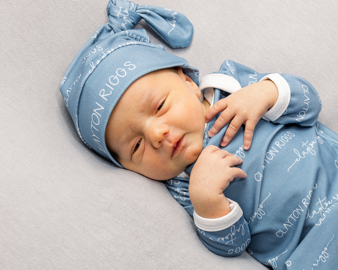A newborn baby wearing a blue onesie with the name "CLAYTON RIGGS" printed on it, lying on a gray surface.