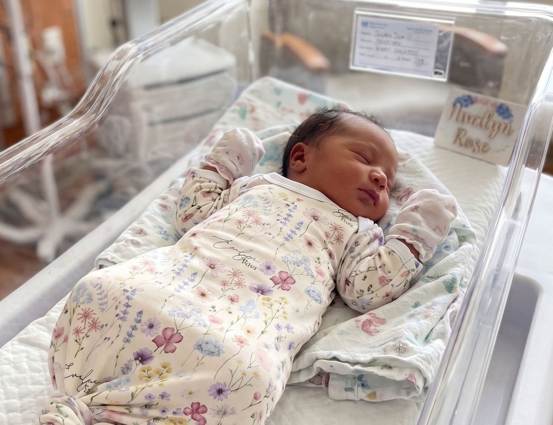 A newborn baby girl is peacefully sleeping in a hospital bed, wearing a white onesie with a floral pattern.