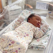 A newborn baby girl is peacefully sleeping in a hospital bed, wearing a white onesie with a floral pattern.