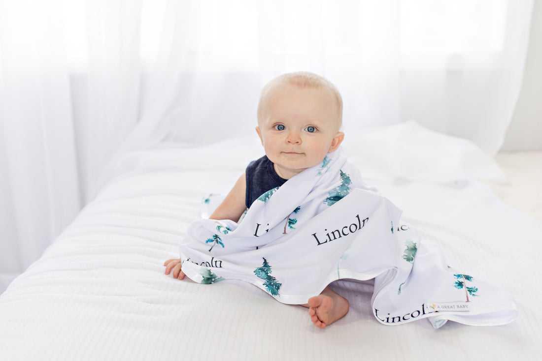A baby is sitting on a bed, wrapped in a blanket with the word "Lincoln" printed on it.