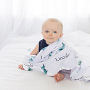 A baby is sitting on a bed, wrapped in a blanket with the word "Lincoln" printed on it.