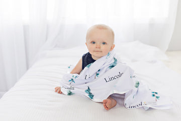 A baby is sitting on a bed, wrapped in a blanket with the word "Lincoln" printed on it.