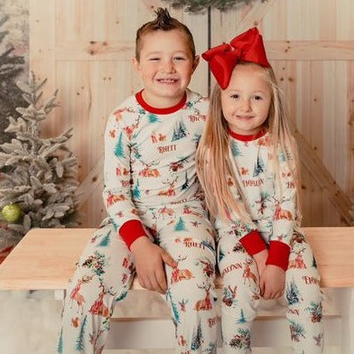 little kids wearing Christmas in the Forest soft and stretchy Christmas Pajamas with forest animals, trees, and pinecone bundles decorated for the holidays, on white fabric and red cuffs, with child's name scattered among the images.