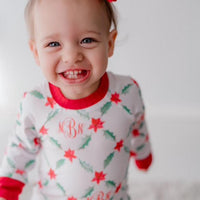 LIttle girl wearing personalized Holiday Pajamas for Girls with coquette style lattice holly leaf print, red ribbins, and initials monogrammed in red, on white jersey knit background and red cuffs.