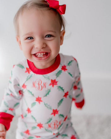 LIttle girl wearing personalized Holiday Pajamas for Girls with coquette style lattice holly leaf print, red ribbins, and initials monogrammed in red, on white jersey knit background and red cuffs.