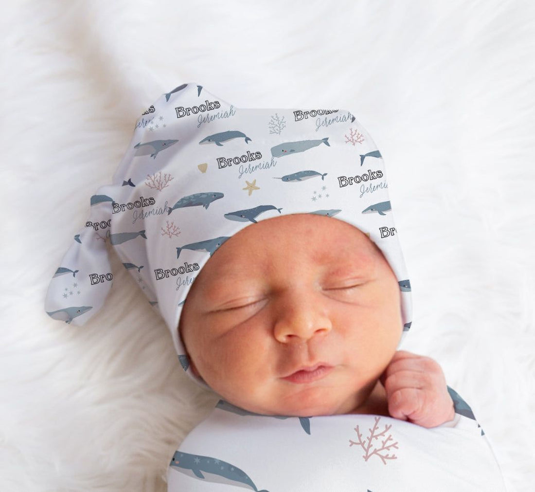 A sleeping newborn baby swaddled in a white blanket with a pattern of blue and gray sea creatures, wearing a hat with the name "Brooks" printed on it.