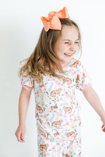 little girl wearing classic Easter bunny pajamas, Girl's custom aster pajamas with brown bunnies, and pink and yellow flowers scattered around white knit fabric along with her name, pale pink cuffs. 