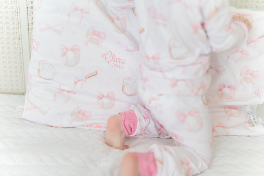 little girl laying on a personalized minky pillowcase with pink bows and baseball equipment on a pale pink background and girl's name. 