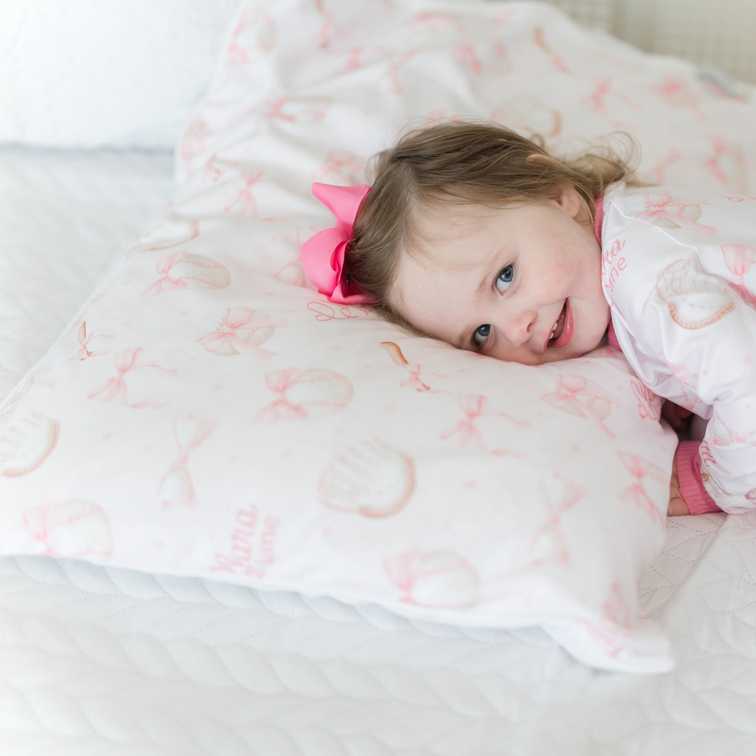 little girl laying on a personalized minky pillowcase with pink bows and baseball equipment on a pale pink background and girl's name. 