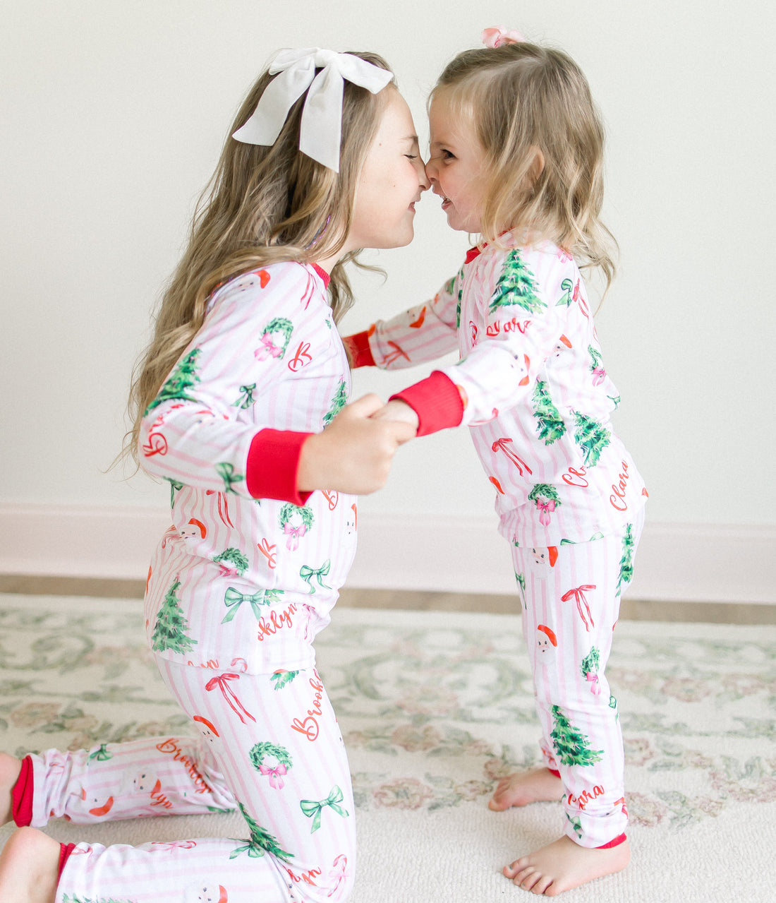 Two children in matching personalized pajamas with Christmas tree and pink stripe patterns, standing close together on a light wooden floor.