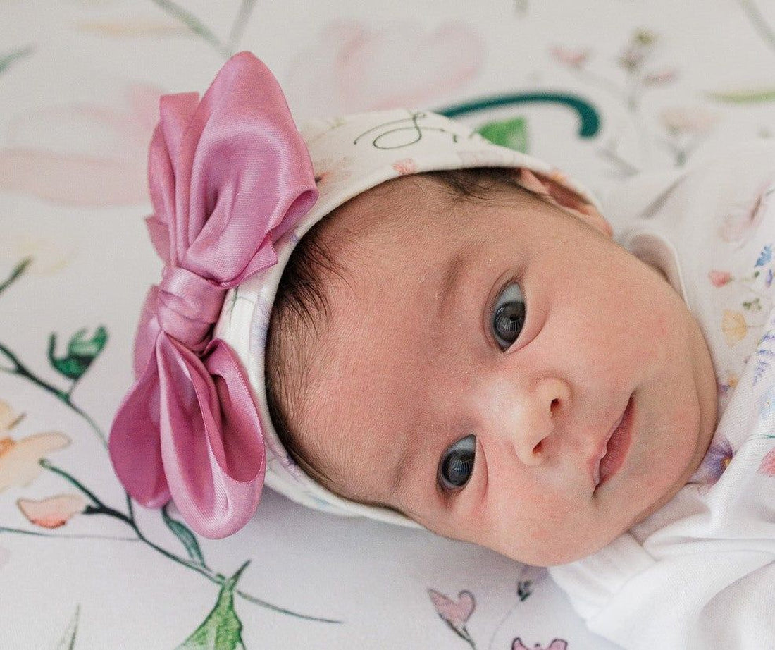 A close-up portrait of a baby girl wearing a pink bow on her head, lying on a floral-patterned blanket.
