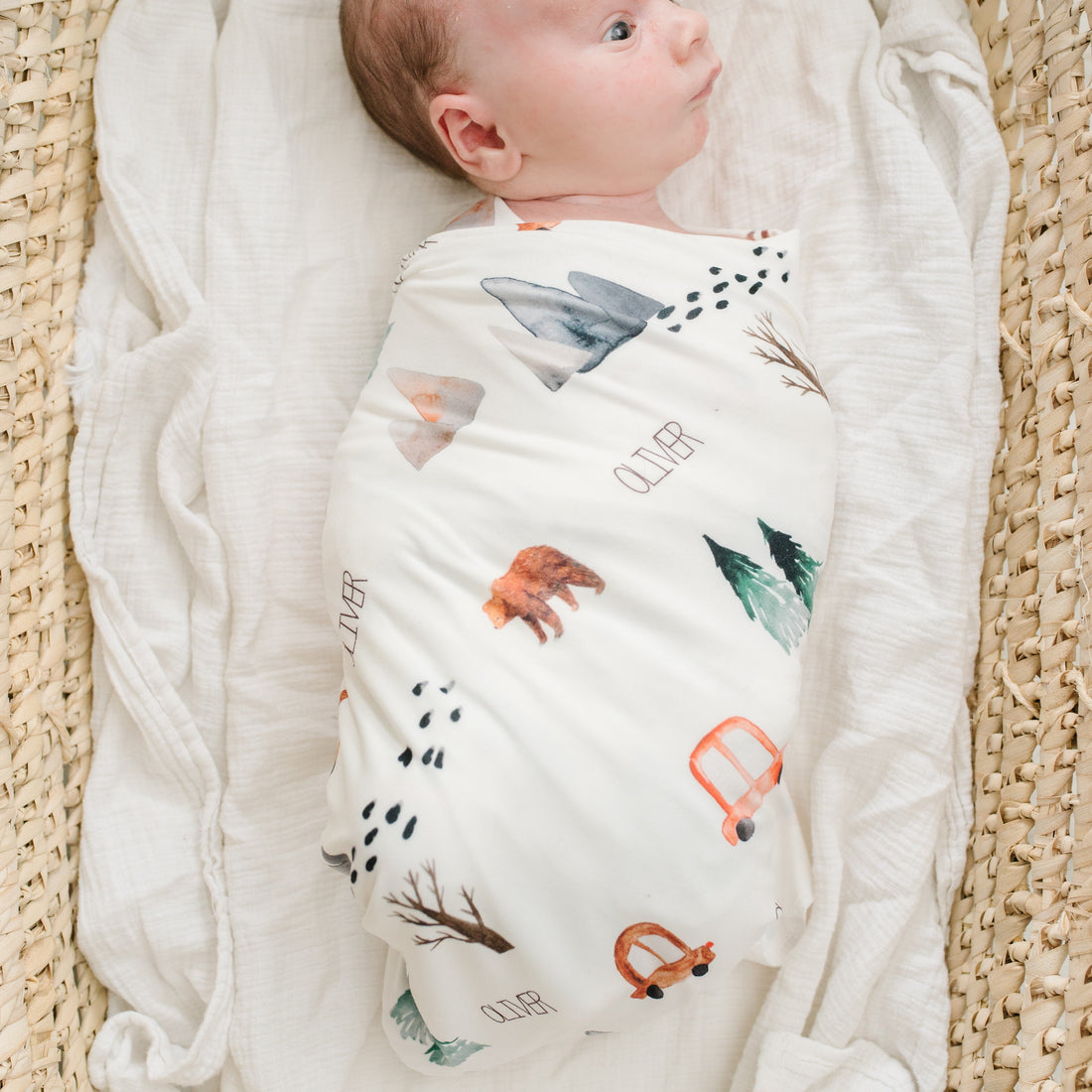 A baby swaddled in a white blanket with various animal and vehicle designs, lying in a woven basket.