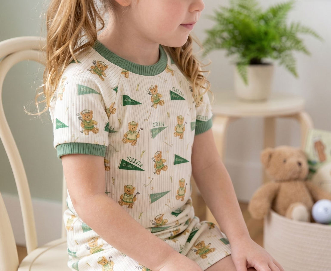 A young girl wearing a white and green pajama top with a pattern of teddy bears is sitting on a chair.