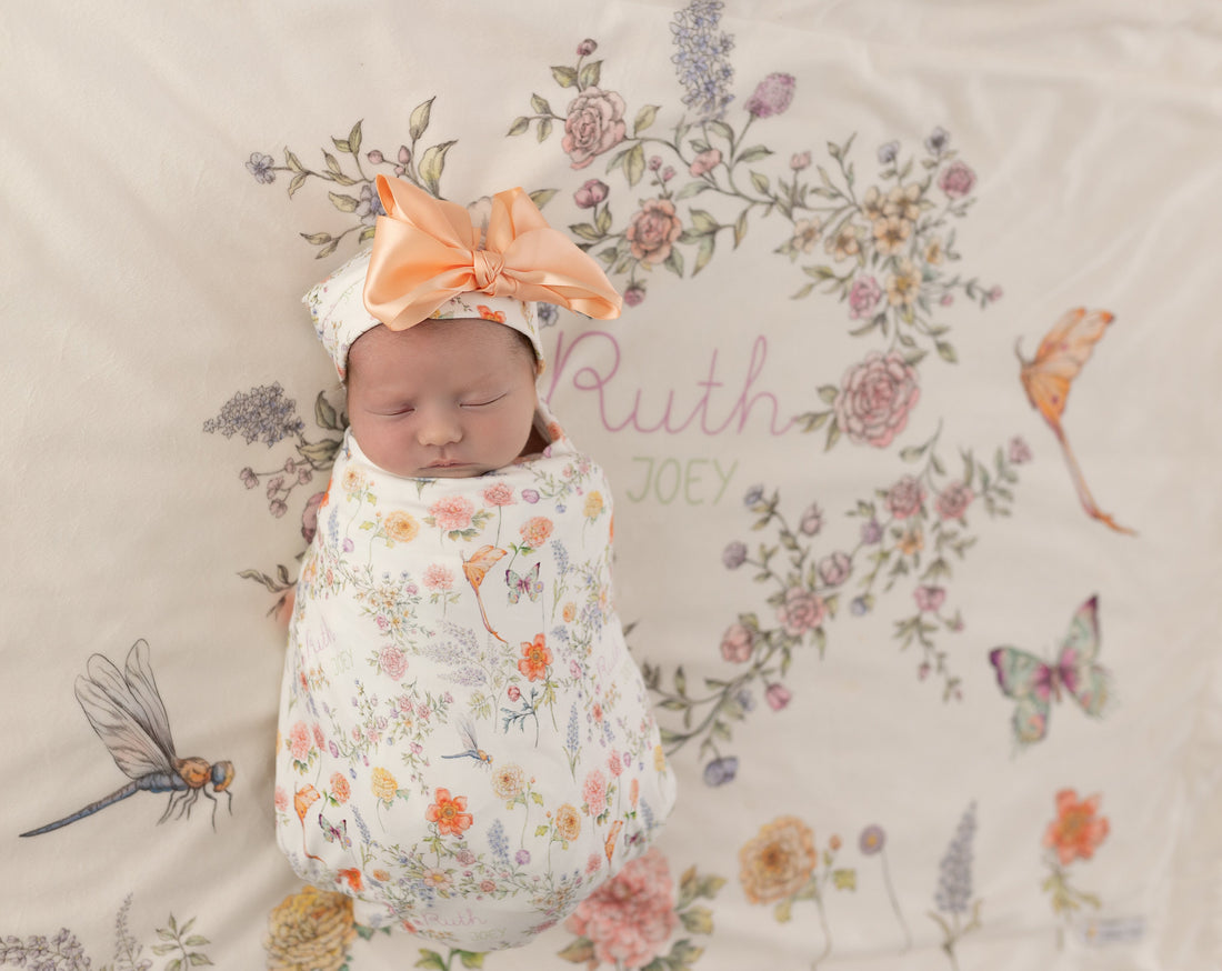 A newborn baby girl is peacefully sleeping, swaddled in a white blanket with a floral pattern and wearing a matching headband. The baby's name, "Ruth", is written on the blanket, and the blanket also features a butterfly and a dragonfly design.