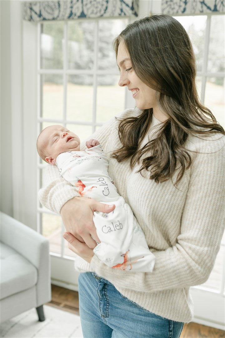 A woman in a white sweater holds a sleeping newborn baby wrapped in a blanket with the name "Caleb" printed on it.