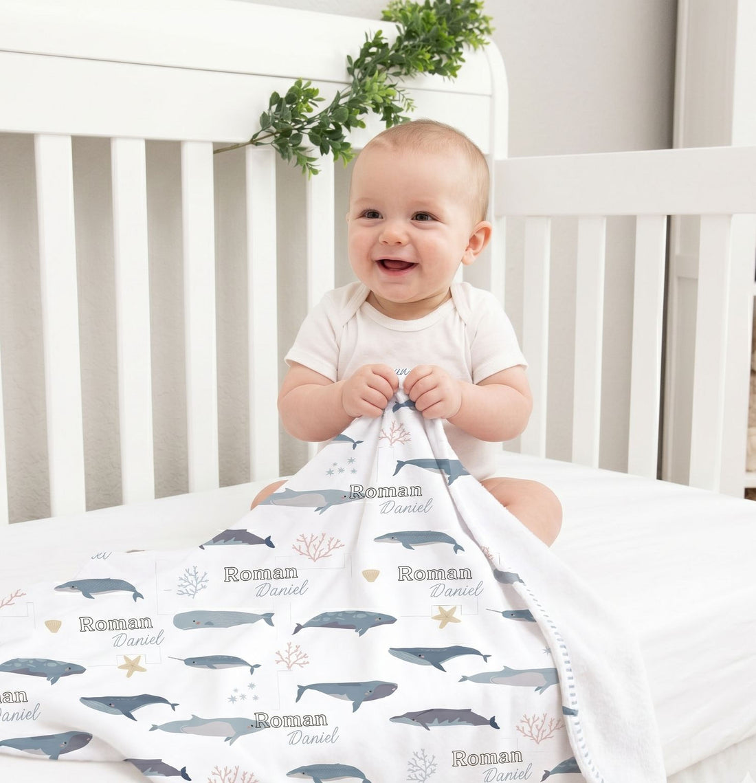A smiling baby boy sitting in a white crib, holding a blanket with a whale and starfish pattern.