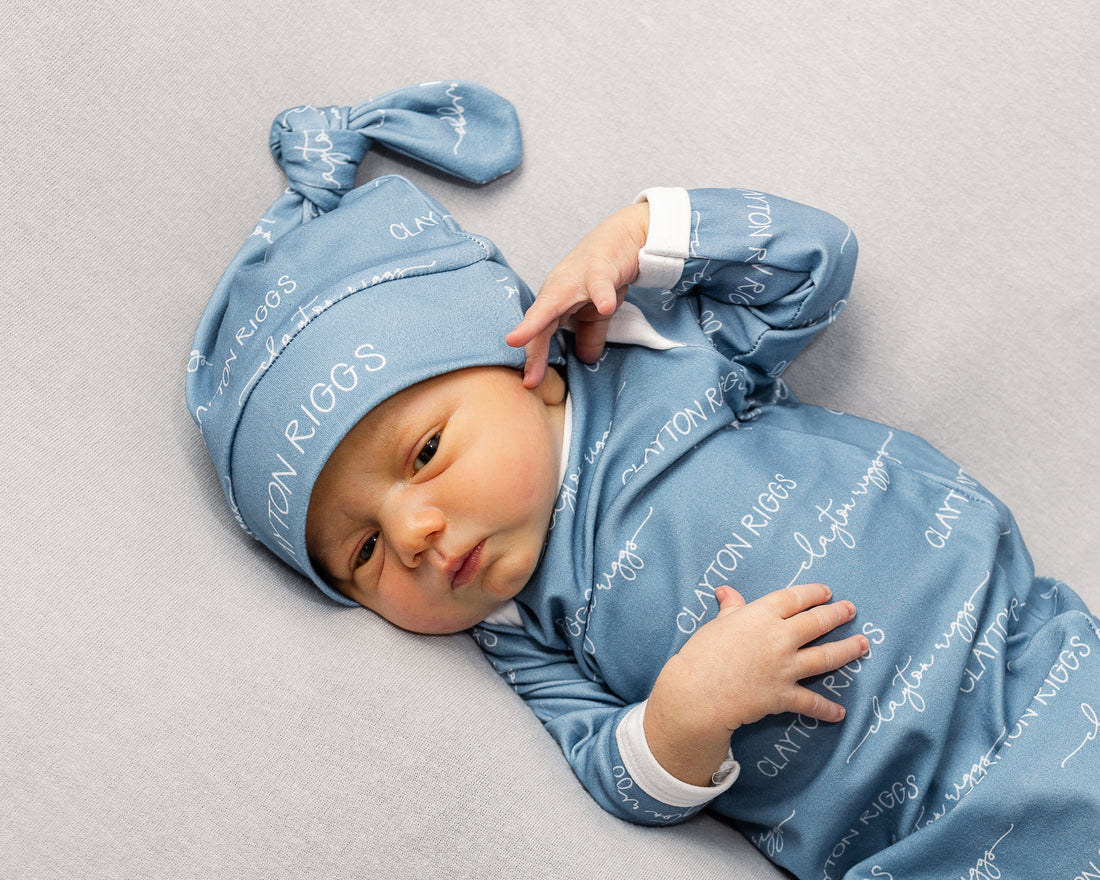 A newborn baby wearing a blue onesie with the word "CLAYTON RIGGS" printed on it, lying on a gray surface.
