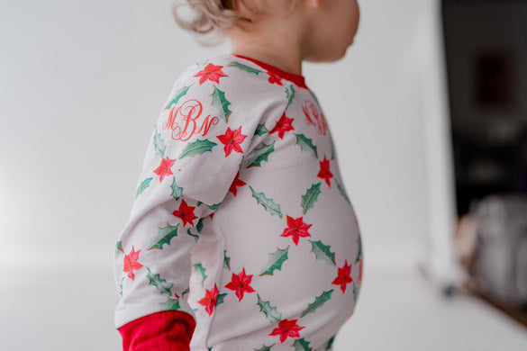 LIttle girl wearing personalized Holiday Pajamas for Girls with coquette style lattice holly leaf print, red ribbins, and initials monogrammed in red, on white jersey knit background and red cuffs.