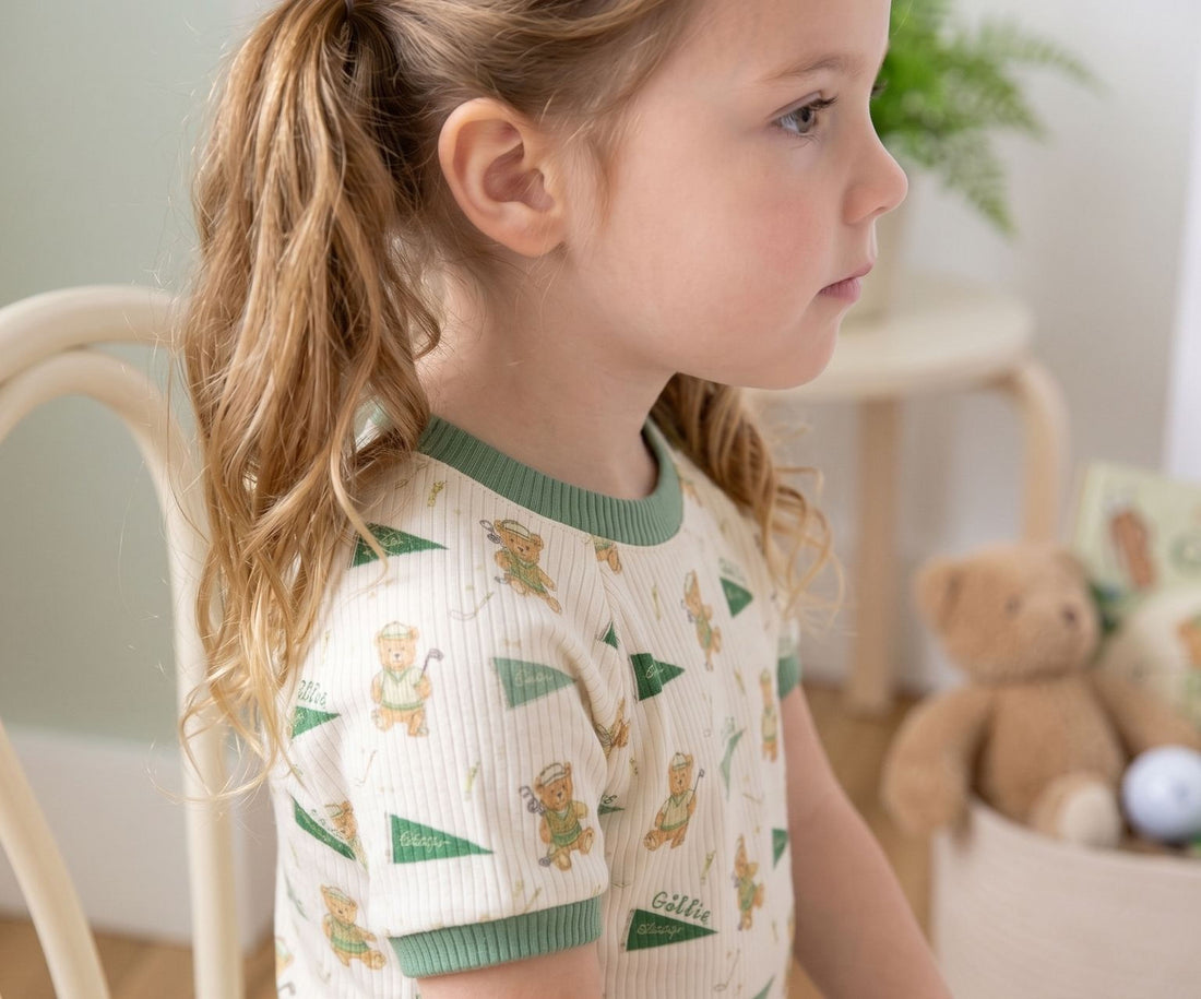 A young girl with blonde hair is sitting in a chair, wearing a white shirt with green and orange teddy bear designs.