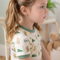 A young girl with blonde hair is sitting in a chair, wearing a white shirt with green and orange teddy bear designs.