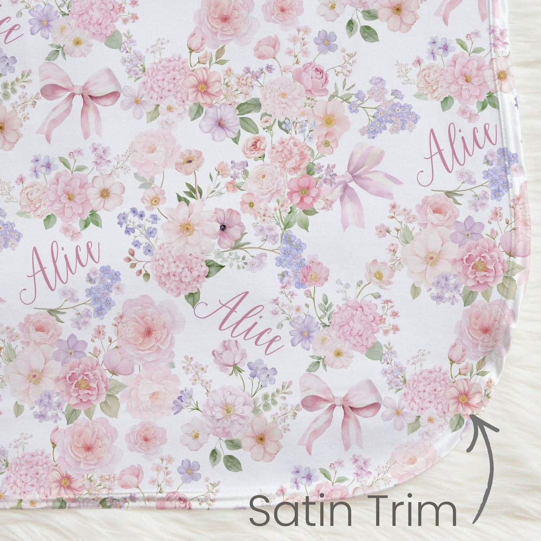a floral print blanket with pink and purple flowers, bows, and the name "Alice" written on it. The blanket has a satin trim, and there is a small text label indicating it is a satin trim.