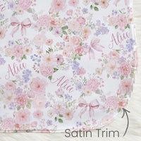 a floral print blanket with pink and purple flowers, bows, and the name "Alice" written on it. The blanket has a satin trim, and there is a small text label indicating it is a satin trim.