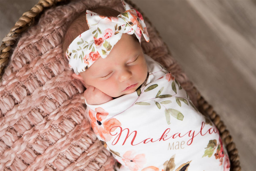 A newborn baby girl is peacefully sleeping in a basket, wearing a white floral headband and a blanket with the name "Makayla" printed on it.