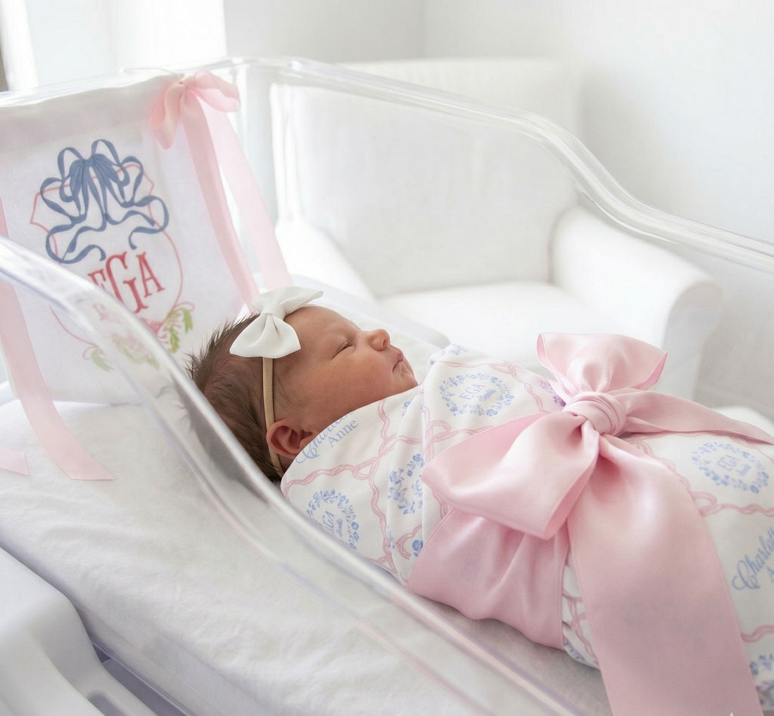 A newborn baby girl is peacefully sleeping in a hospital bed, wearing a pink bow and a white headband.