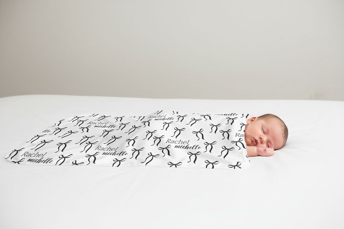 A newborn baby is peacefully sleeping on a bed, wrapped in a blanket with the name "Rachael" and "Michael" printed on it.