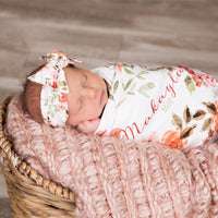 A newborn baby girl is peacefully sleeping in a basket, wrapped in a floral blanket and wearing a white headband with a bow.