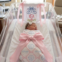 A newborn baby girl is peacefully sleeping in a hospital bed, wrapped in a pink blanket with a large pink ribbon.